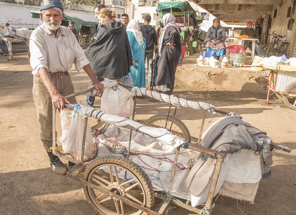 white-bearded man with handmade cart offers transport services to outdoor market customers.